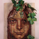 an intense green and brown face trapped in a cage staring out at the audience; the cage is partially broken open along a seam, due to the weathering caused by a large grape vine that has grown on the top of the cage, large dark and light green aluminum can grape leaves and grapes made of found objects are adhered in a light, scattered, cascading fashion across the top and right side of the cage