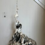 a sculpture of a figure appearing to emerge out of a thick, batter-like substance, dark at the base with many layers of poetry text, gradually becoming lighter and less dense as you look up toward the top of the sculpture that is creamy white with no text at all; the figure is twisted and contorted as though tired and struggling and the arm is dramatically upstretched in a straight long, thin, antenna-like form with a tiny grasping hand at the top; the poem title Questions of "I" wraps clearly around the arm; layers of the thick batter drip down and peel back away from the antenna arm; the face is only a concave mirror facing upward like an eager, empty tray waiting to be filled