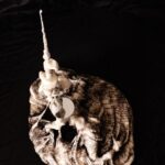 a sculpture of a figure appearing to emerge out of a thick, batter-like substance, dark at the base with many layers of poetry text, gradually becoming lighter and less dense as you look up toward the top of the sculpture that is creamy white with no text at all; the figure is twisted and contorted as though tired and struggling and the arm is dramatically upstretched in a straight long, thin, antenna-like form with a tiny grasping hand at the top; the poem title Questions of "I" wraps clearly around the arm; layers of the thick batter drip down and peel back away from the antenna arm; the face is only a concave mirror facing upward like an eager, empty tray waiting to be filled