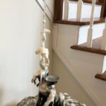 a sculpture of a figure appearing to emerge out of a thick, batter-like substance, dark at the base with many layers of poetry text, gradually becoming lighter and less dense as you look up toward the top of the sculpture that is creamy white with no text at all; the figure is twisted and contorted as though tired and struggling and the arm is dramatically upstretched in a straight long, thin, antenna-like form with a tiny grasping hand at the top; the poem title Questions of "I" wraps clearly around the arm; layers of the thick batter drip down and peel back away from the antenna arm; the face is only a concave mirror facing upward like an eager, empty tray waiting to be filled