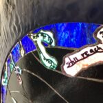 a collection of different sized, hands all in different positions with tree-bark like coloring and texturing are in a silhouetted grassy brambly field wiht sunset colors behind in the sky; the hands are protruding inward from a square substrate and encircling a stained glass circular image of the sky with the Aurora Borealis and moon at the center; the fingertips of the hands turn into tree branches that have lines of poetry resting on them; where the branches reach the glass sky, they become the poetry coated in colorful ice as though a tree after an ice storm, being lit by the Northern Lights