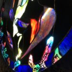 a collection of different sized, hands all in different positions with tree-bark like coloring and texturing are in a silhouetted grassy brambly field wiht sunset colors behind in the sky; the hands are protruding inward from a square substrate and encircling a stained glass circular image of the sky with the Aurora Borealis and moon at the center; the fingertips of the hands turn into tree branches that have lines of poetry resting on them; where the branches reach the glass sky, they become the poetry coated in colorful ice as though a tree after an ice storm, being lit by the Northern Lights