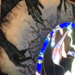 a collection of different sized, hands all in different positions with tree-bark like coloring and texturing are in a silhouetted grassy brambly field wiht sunset colors behind in the sky; the hands are protruding inward from a square substrate and encircling a stained glass circular image of the sky with the Aurora Borealis and moon at the center; the fingertips of the hands turn into tree branches that have lines of poetry resting on them; where the branches reach the glass sky, they become the poetry coated in colorful ice as though a tree after an ice storm, being lit by the Northern Lights