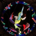 a collection of different sized, hands all in different positions with tree-bark like coloring and texturing are in a silhouetted grassy brambly field wiht sunset colors behind in the sky; the hands are protruding inward from a square substrate and encircling a stained glass circular image of the sky with the Aurora Borealis and moon at the center; the fingertips of the hands turn into tree branches that have lines of poetry resting on them; where the branches reach the glass sky, they become the poetry coated in colorful ice as though a tree after an ice storm, being lit by the Northern Lights