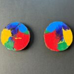 Two rustic round live-edge coasters with mirror image copies of the same non-representational radial image with acrylic swaths of yellow, light blue, red, green, and dark blue at the center