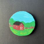 Rustic round live-edge coaster with a red barn in a field with a line of trees behind painted on the surface