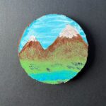 Rustic round live-edge coaster with a snowcapped mountain range with streaky clouds above and a green valley and river below painted on the surface