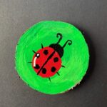 Rustic round live-edge coaster with a shiny ladybug that has curly antennas against a bright green background painted on the surface