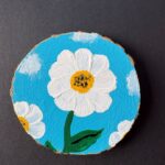 Rustic round live-edge coaster with a bright white daisy set against a bright blue sky with some clouds painted on the surface