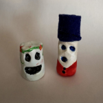 Two sculptures that resemble marshmallows with black indentations for two eyes and expressive mouths; on the left the character has a headband of pink flowers and stems; the character on the right is wearing a tall dark blue felt. billed hat and red shirt with black buttons