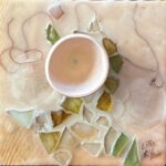 a square sculptural painting hangs from a braided hemp twine with beads; the mixed media painting is beige and cream with swirling lines of brown thread and a figural swoop of mosaic-like cracked glass tiles of green and ochre are embedded in the encaustic; a small bisqueware vessel is fastened just above center