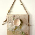 a square sculptural painting hangs from a braided hemp twine with beads; the mixed media painting is beige and cream with swirling lines of brown thread and a figural swoop of mosaic-like cracked glass tiles of green and ochre are embedded in the encaustic; a small bisqueware vessel is fastened just above center