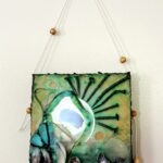 a square low-relief mixed media encaustic painting of cream, turquoise, green and ochre with the iridescent purple of muscle shells that are oriented vertically across the bottom from left to right gradually laying horizontally and appearing to float away; two pieces of similarly sized turquoise glass shards are arranged above the shells and outlined in black that mimic the black of a ray-like, radial arrangement of bent rusty nails arranged small to large and back to small around the top right edge of a circular mirror embedded slightly off-center into the teal-black encaustic medium of the bottom; there are two concave circular scoops out of the wax - one at the top left, the other down to the right of the mirror - revealing the golden wood below
