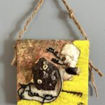 a square low-relief mixed media painting divided in half by a collection of found beach objects each with a notable pair of holes, as well as black leather cord and black text reading "me," "me," "Then you"; the background of the top left is gold and copper leaf and of the bottom right is bright yellow