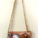 a square mixed media painting of cream, gold, silver, and burgundy that has various circular found objects layered over top of each other, two are mirrors, smaller ones along the edge have holes through the center and some are strung onto the split wire arm of a figure appearing to be turned away from the other arm which is as long as the entire height of the figure and outstretched at a very straight right angle reaching between the two mirrors and appearing to point with its spring-like twisted hand and pointer finger to the underside of a clearly visible golden disc at the top of a heap of less distinguishable discs