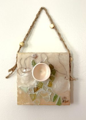 a square sculptural painting hangs from a braided hemp twine with beads; the mixed media painting is beige and cream with swirling lines of brown thread and a figural swoop of mosaic-like cracked glass tiles of green and ochre are embedded in the encaustic; a small bisqueware vessel is fastened just above center